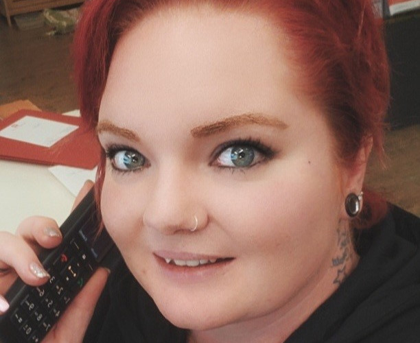 Eine Person mit roten Haaren, Piercings und einem Tattoo am Hals h&auml;lt ein Telefon und l&auml;chelt.